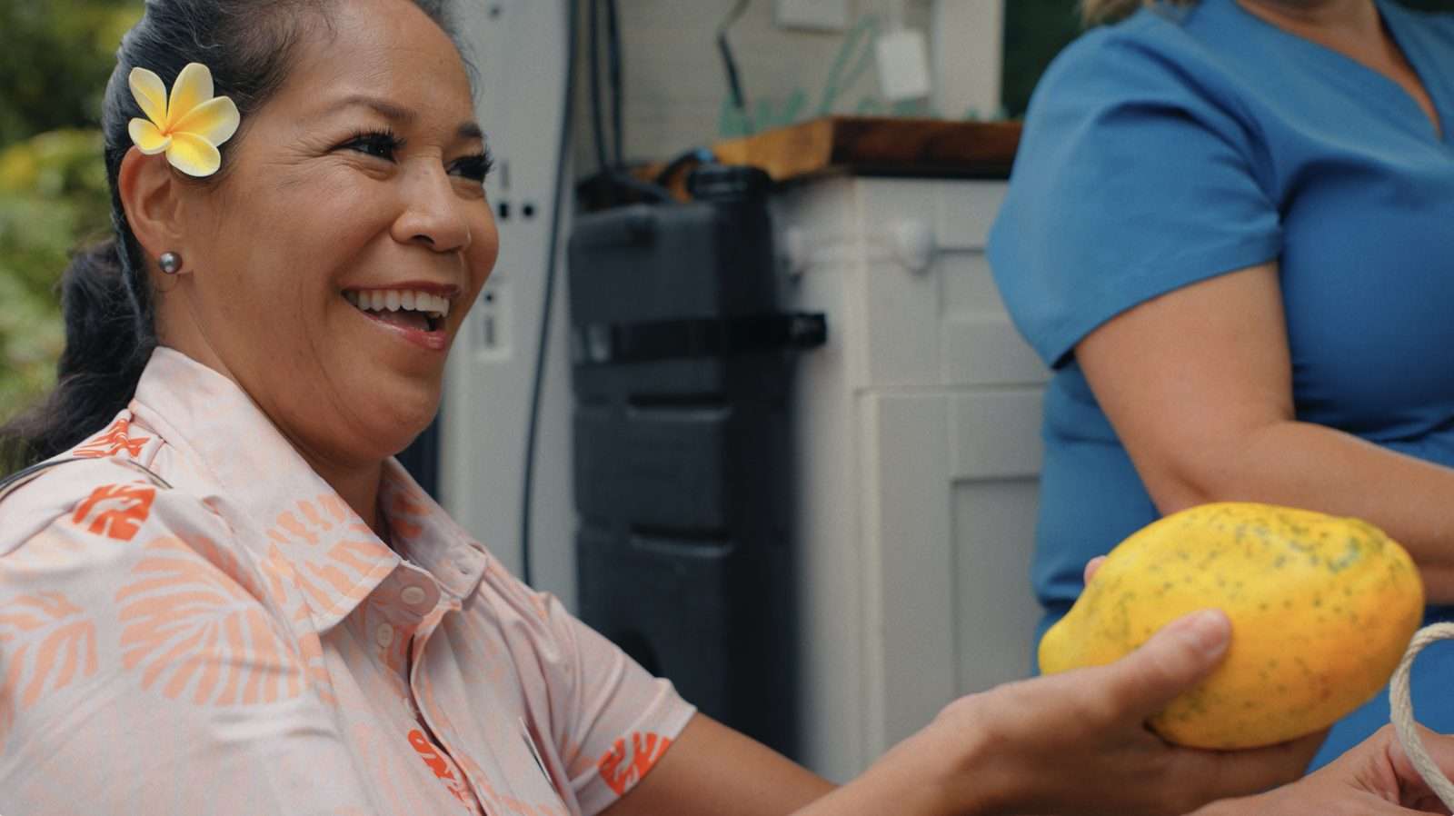 AlohaCare campaign still: a smiling woman with a plumeria in her hair holds out a papaya